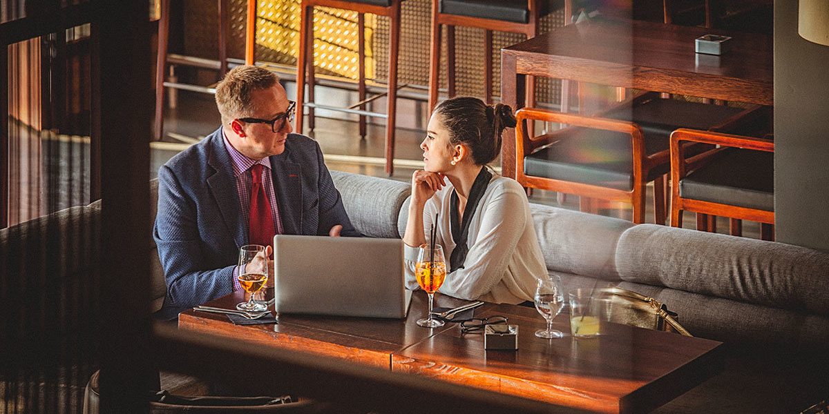 Young business woman and man having a business conversation at a restaurant. They both are well dressed and in a good mood. There is an open laptop on the table before them. Selective focus, waist up, copy space has been left.